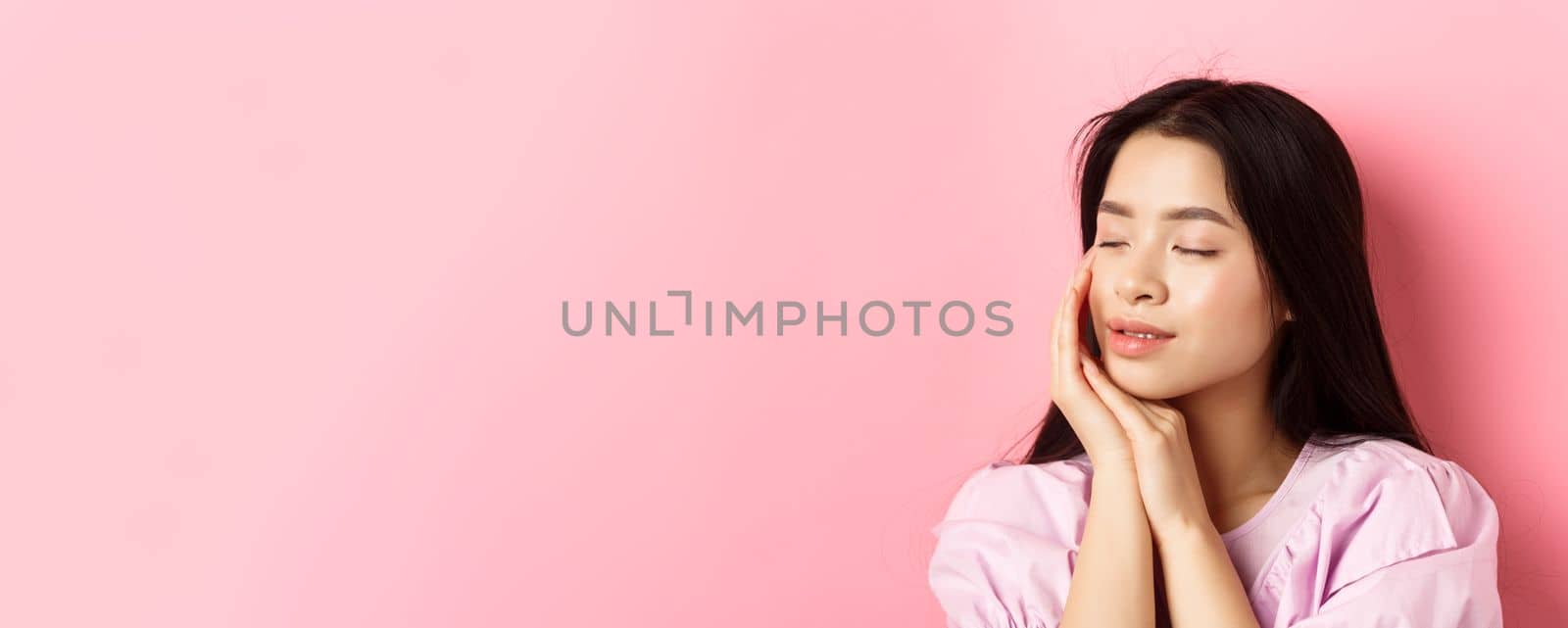 Tender and romantic girl gently touching face, close eyes and smiling softly, standing on pink background. Concept of skincare.