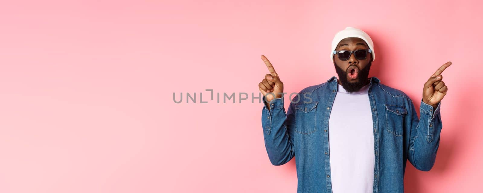 Amazed cool Black guy showing two variants, pointing fingers sideways and staring at camera impressed, wearing sunglasses, standing over pink background.
