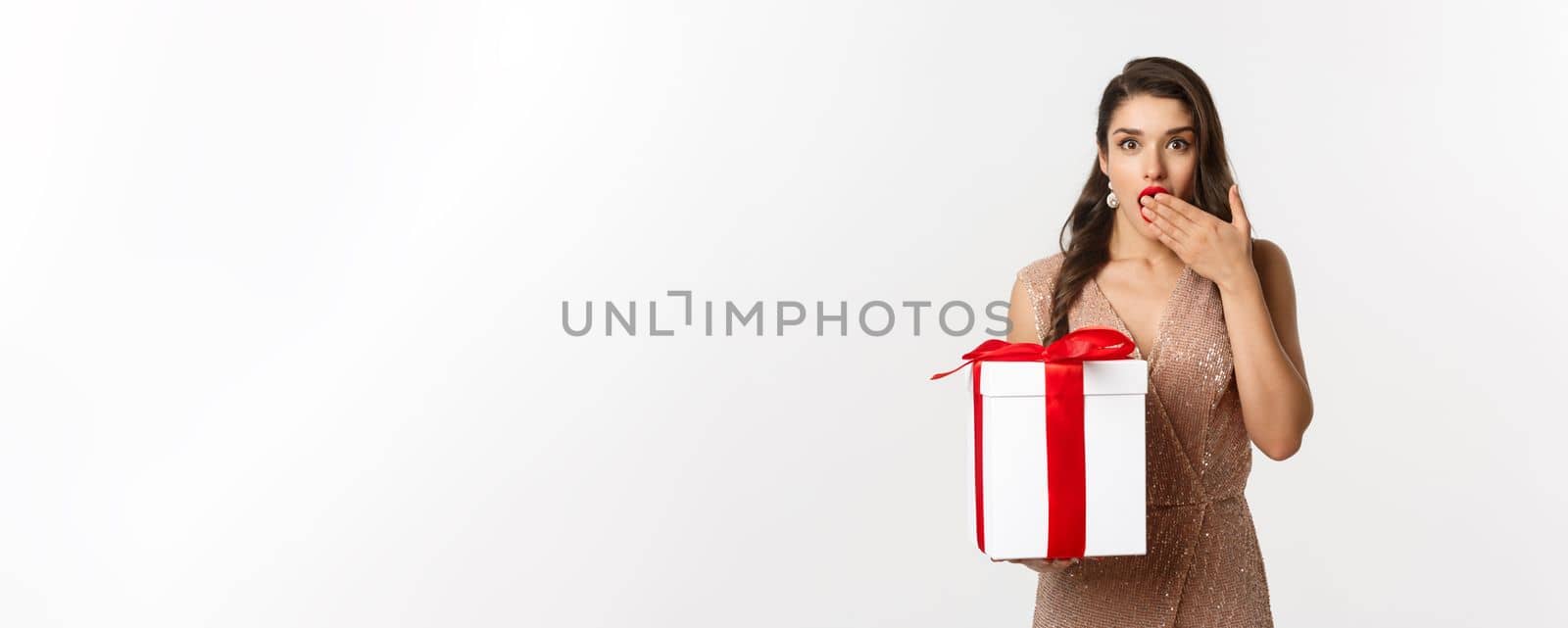Merry Christmas. Beautiful woman looking surprised and holding gift, receiving new year presents, standing in luxury dress, standing over white background.