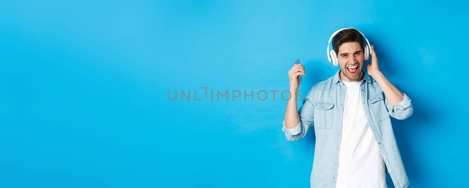 Happy smiling man enjoying listening to music in headphones, holding smartphone in raised hand, standing over blue background.