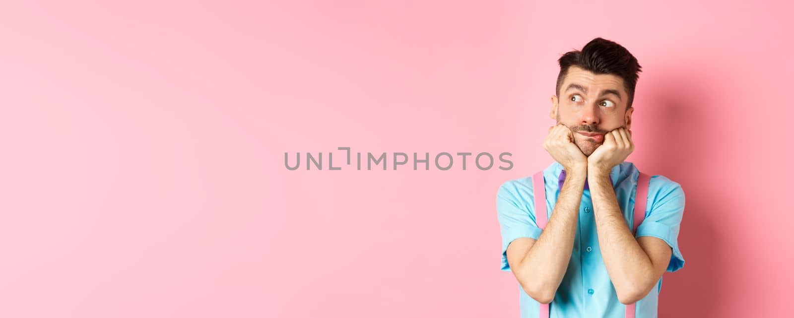 Image of silly and cute young man in suspenders, dreaming of something, looking left with curious face, standing over pink background.