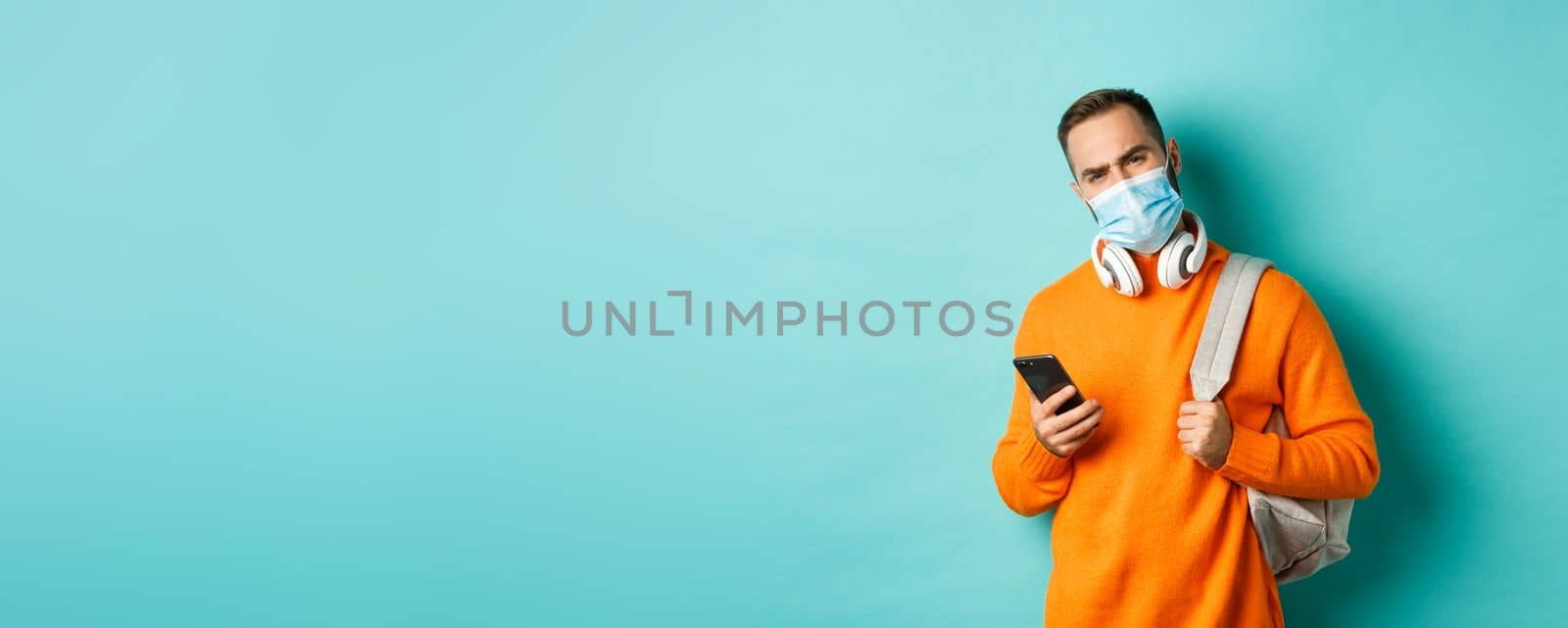 Skeptical and disappointed young man wearing face mask, holding backpack and mobile phone, frowning upset, standing over light blue background.