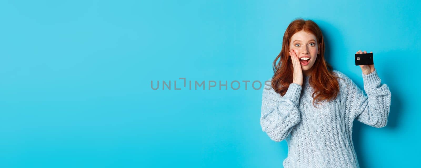 Cute redhead girl in sweater showing credit card, smiling at camera, standing over blue background by Benzoix