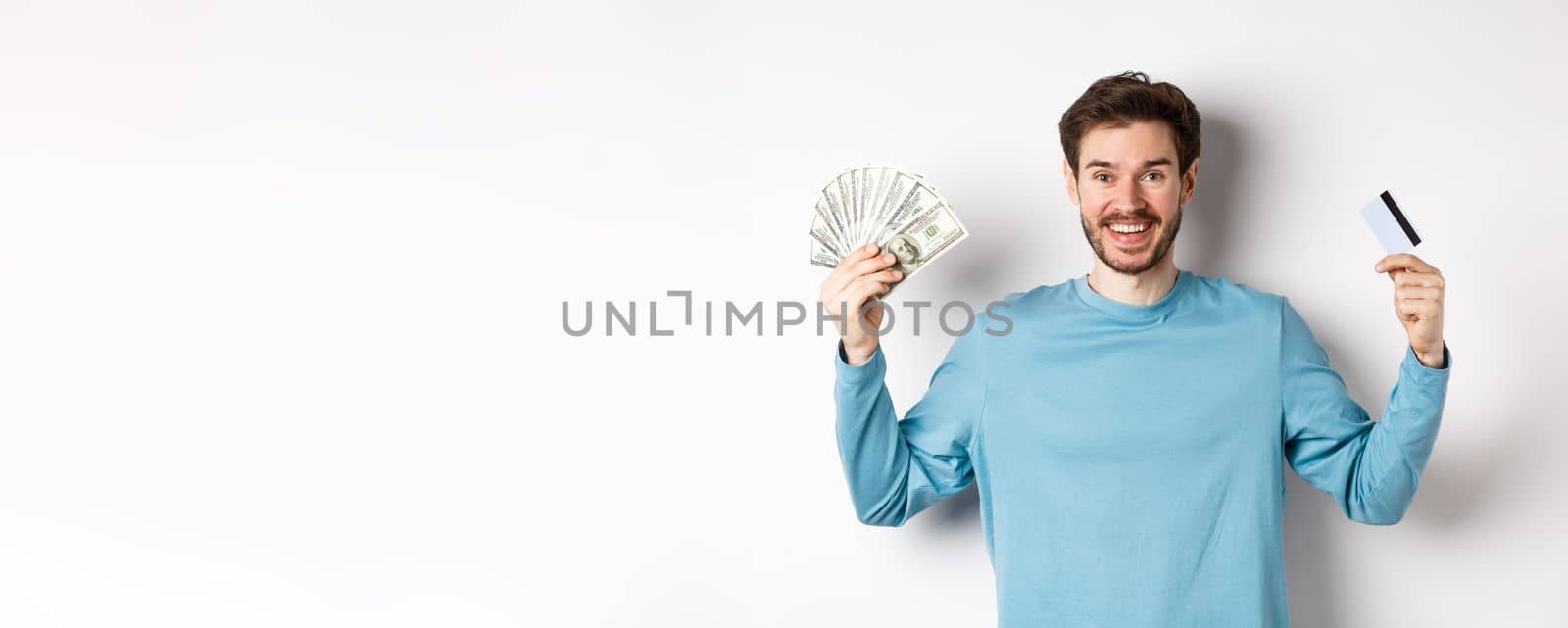 Happy guy smiling and showing plastic credit card with cash, choosing between money and contactless payments, white background by Benzoix