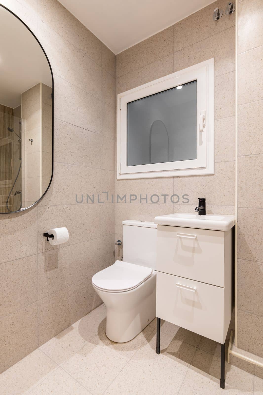 Part of bathroom with toilet bowl, washbasin on table, beautiful oval mirror on wall lined with beige marble tiles, in which you can see reflection of shower area with metal fittings