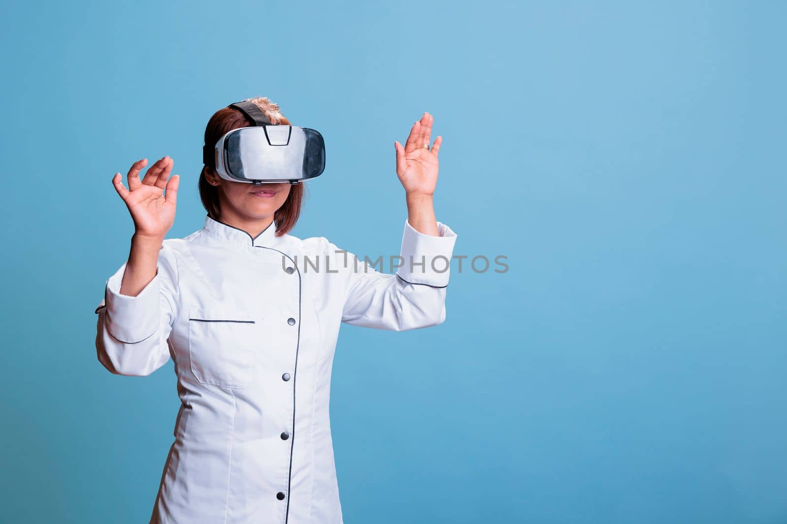 Friendly cook worker using virtual reality goggles while cooking healthy meal in studio with blue background. Asian chef preparing culinary recipe for restaurant. Food industry concept