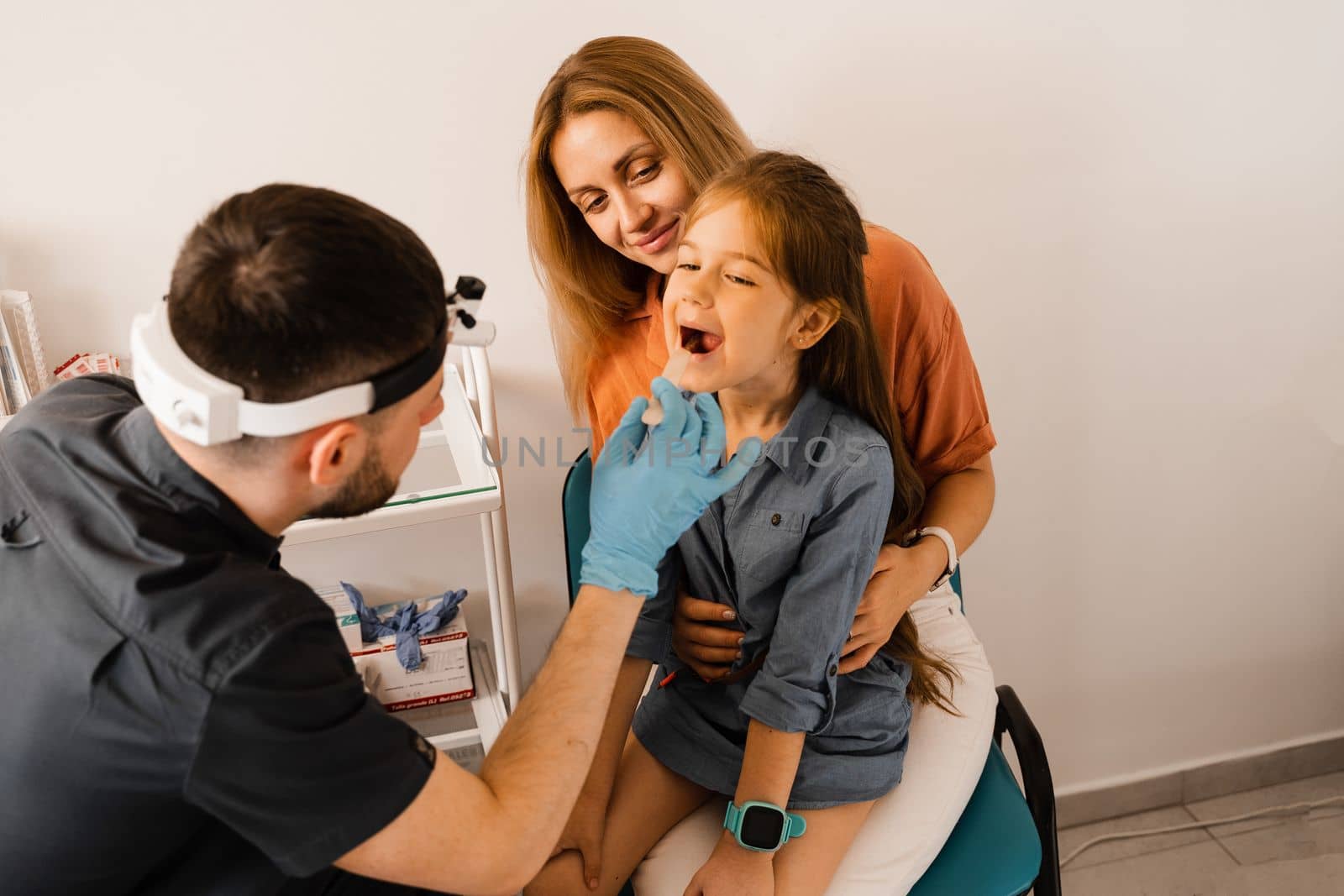 Oropharyngoscopy family procedure for child. Mother with daughter visit ENT doctor. Otolaryngologist examines child throat with spatula. Family consultation with laryngologist. by Rabizo