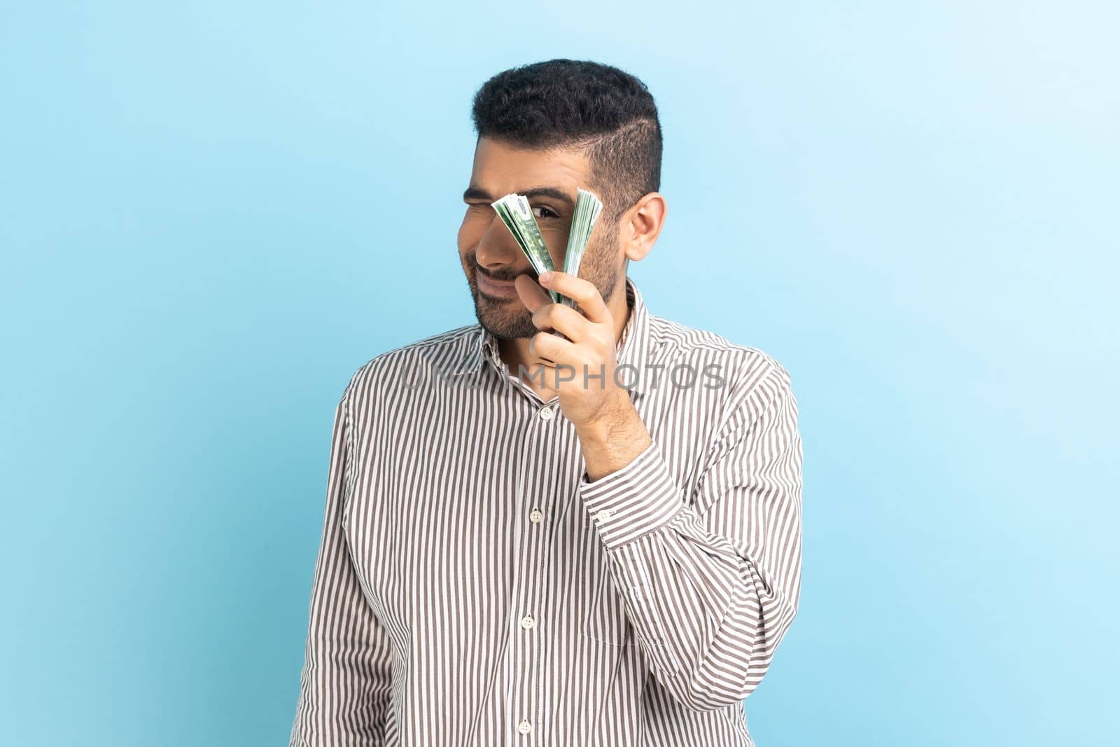 Portrait of curious positive funny businessman peeping through bunch of euro banknotes, passive income, wearing striped shirt. Indoor studio shot isolated on blue background.