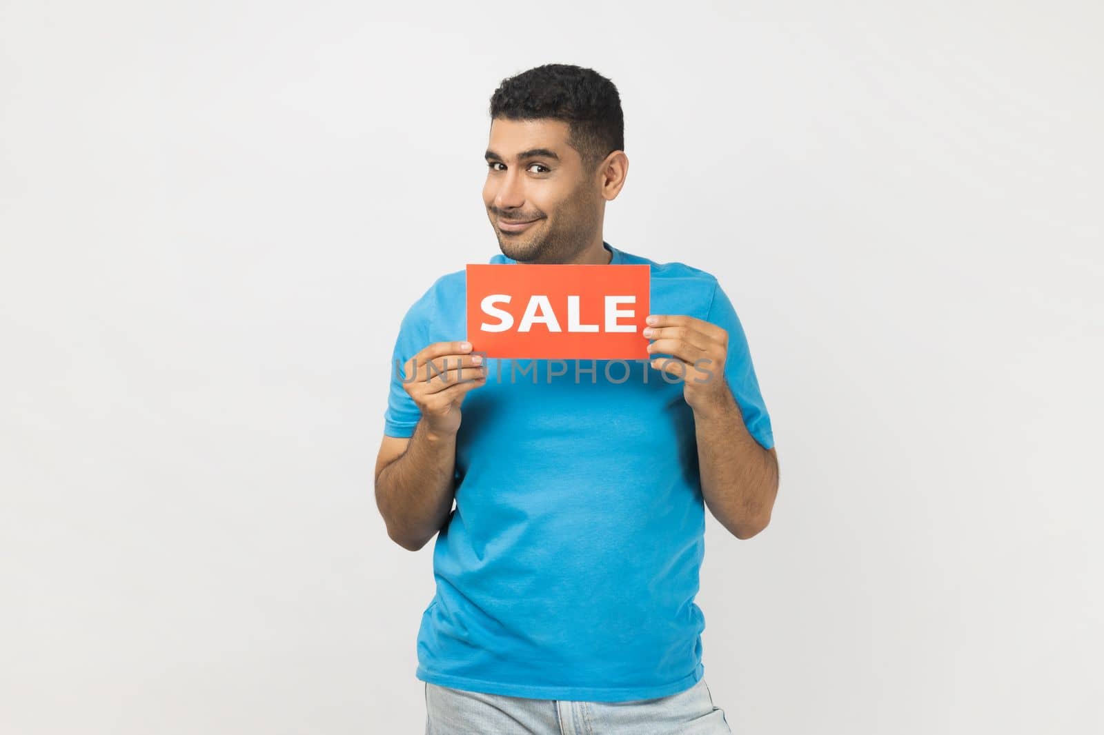 Portrait of positive unshaven man wearing blue T- shirt standing holding sale card in hands looking at camera with toothy smile, black friday. Indoor studio shot isolated on gray background.
