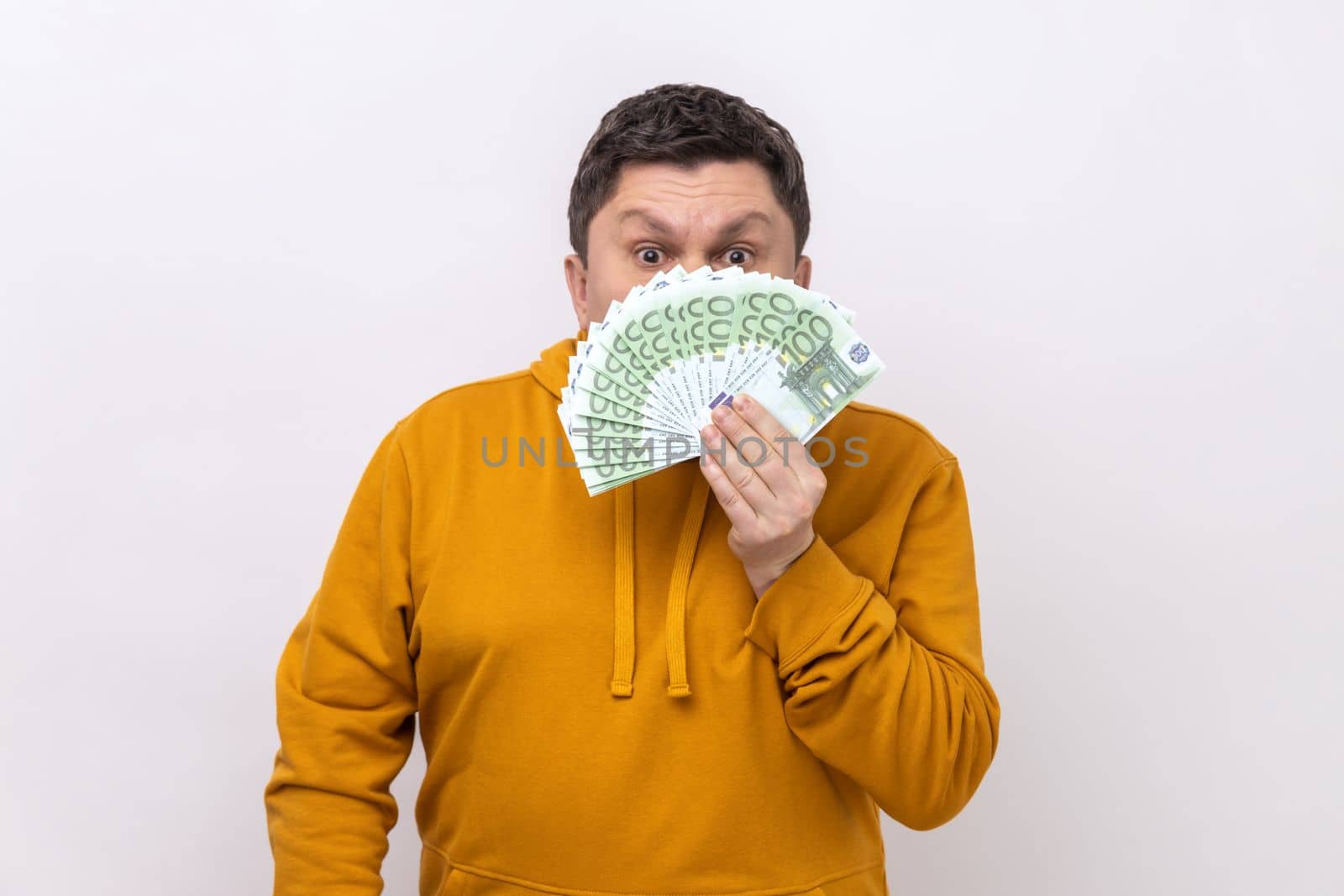 Dark haired man hiding half of face behind fan of hundred euro bills, big winning, betting, wearing urban style hoodie. Indoor studio shot isolated on white background.
