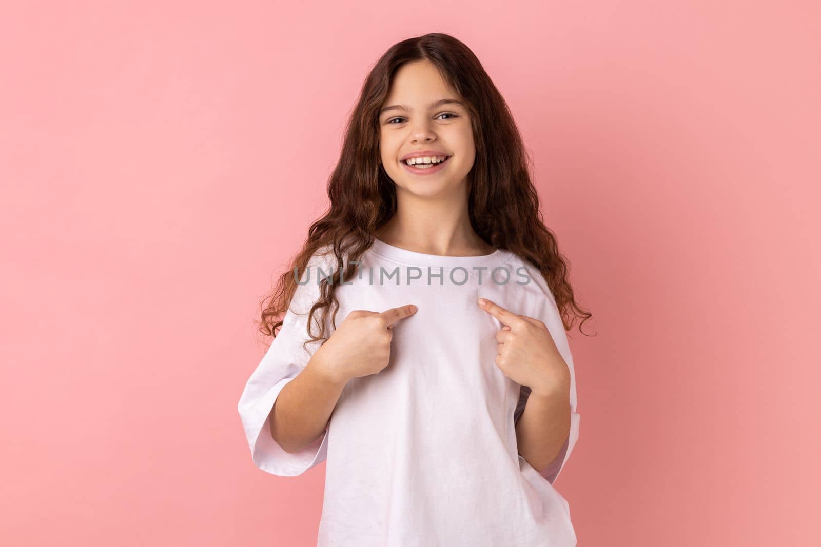 Portrait of ambitious little girl wearing white T-shirt with toothy smile on face pointing fingers on himself, confident teenager. Indoor studio shot isolated on pink background.