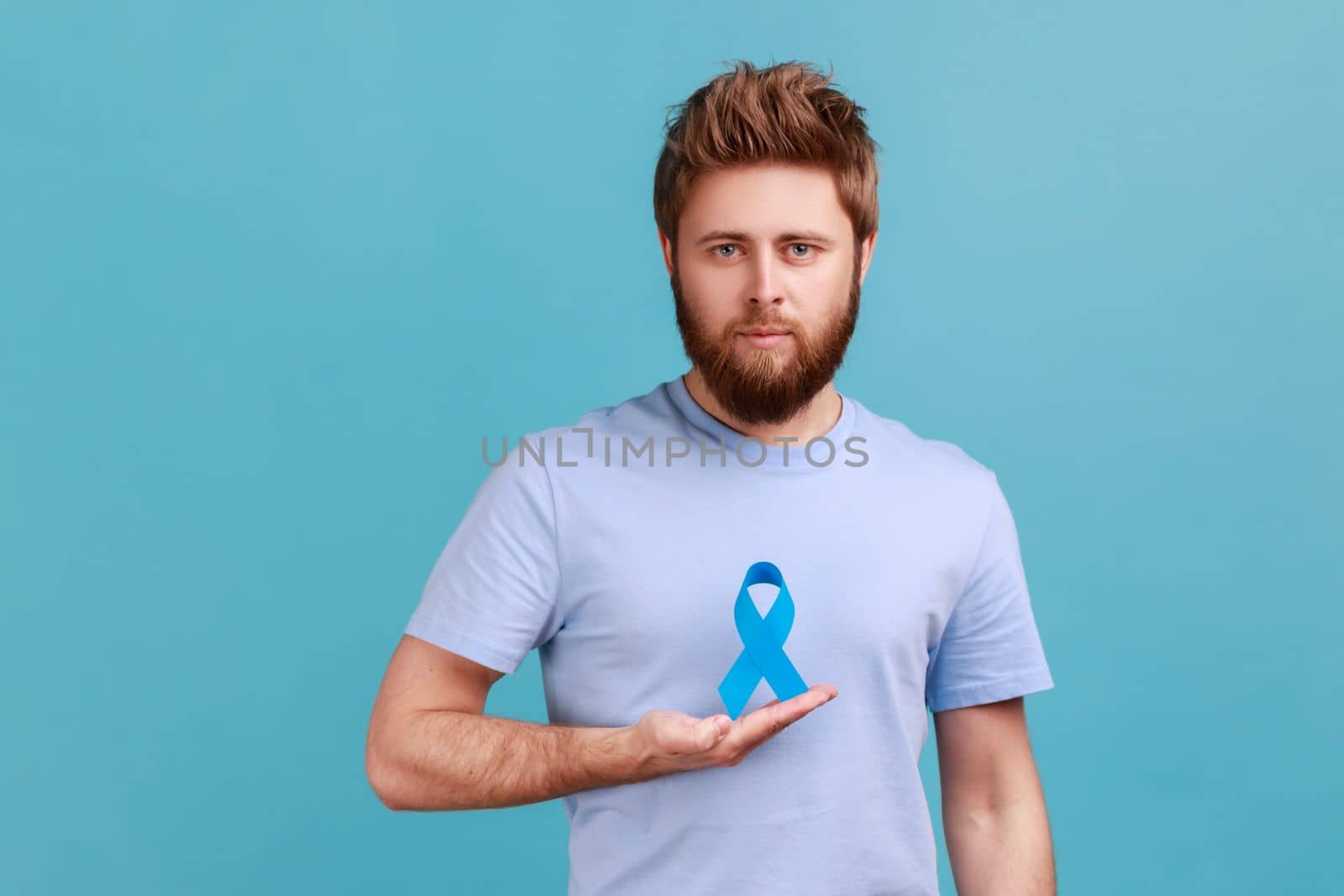 Portrait of serious young adult attractive bearded man holding blue awareness, disease symbol, looking at camera, support and care. Indoor studio shot isolated on blue background.