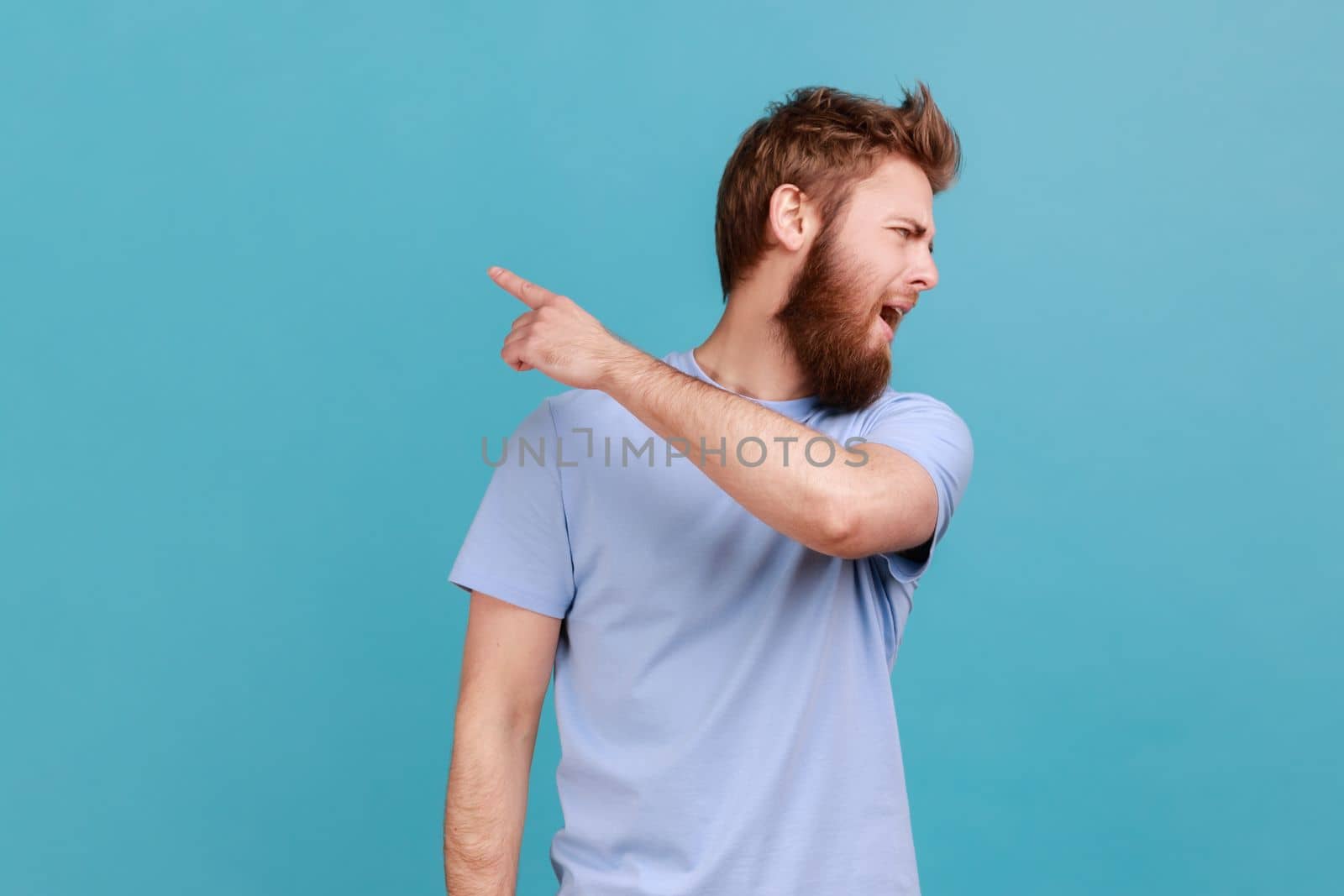 Get out. Portrait of handsome bearded man looking aside and pointing finger another way, asking to leave, dissatisfied expression. Indoor studio shot isolated on blue background.