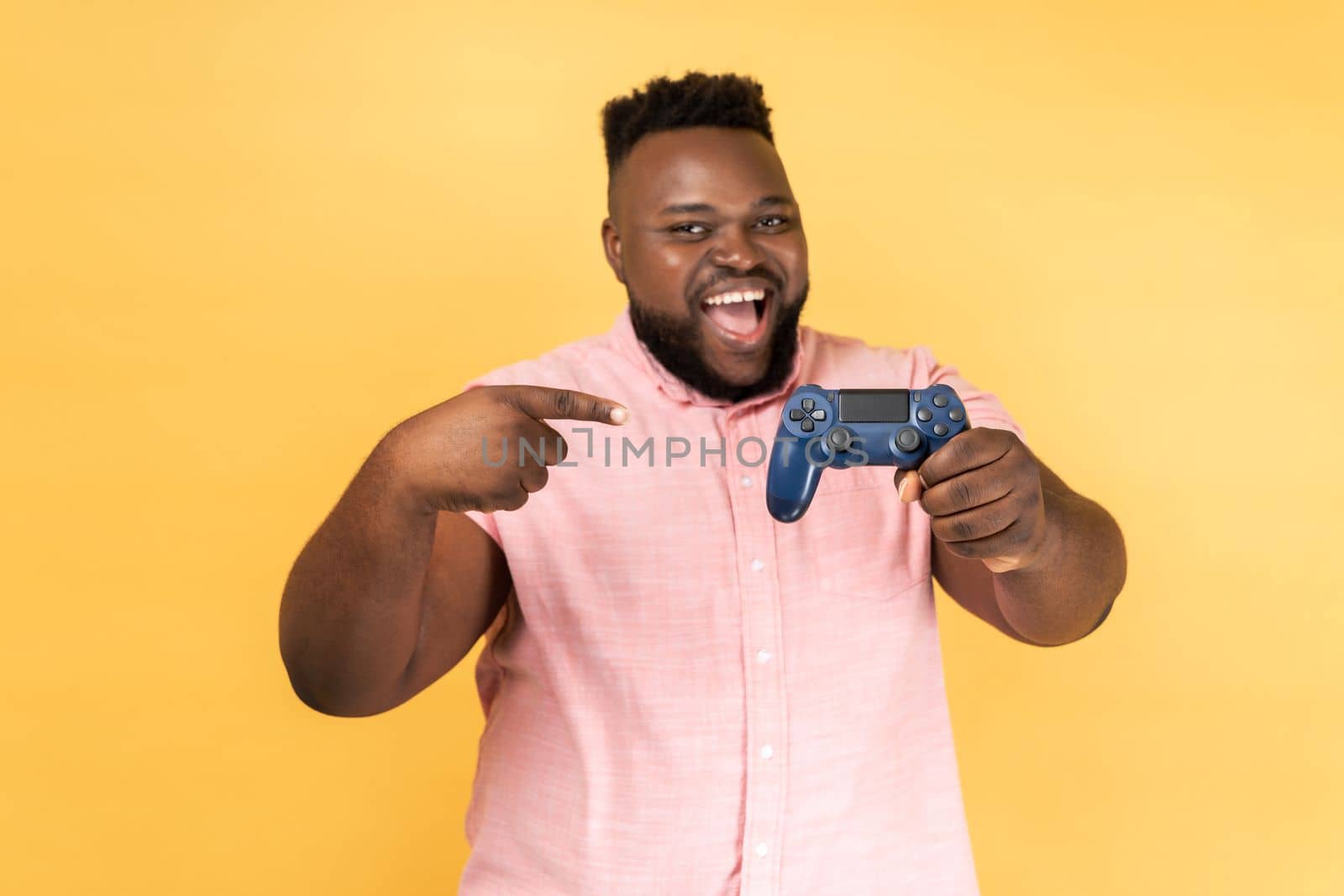 Portrait of excited happy positive bearded man wearing pink shirt standing with excited expression, holding pointing at joystick. Indoor studio shot isolated on yellow background.
