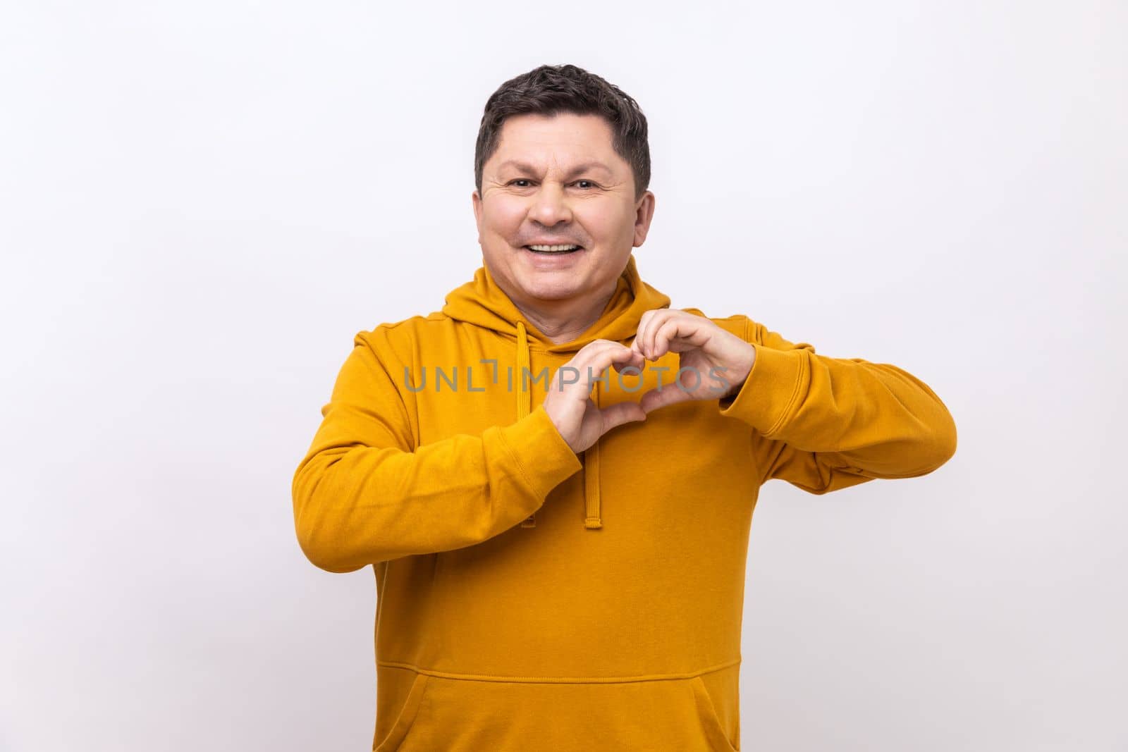 Happy handsome dark haired man standing with heart or love gesture and looking at camera with toothy smile, wearing urban style hoodie. Indoor studio shot isolated on white background.