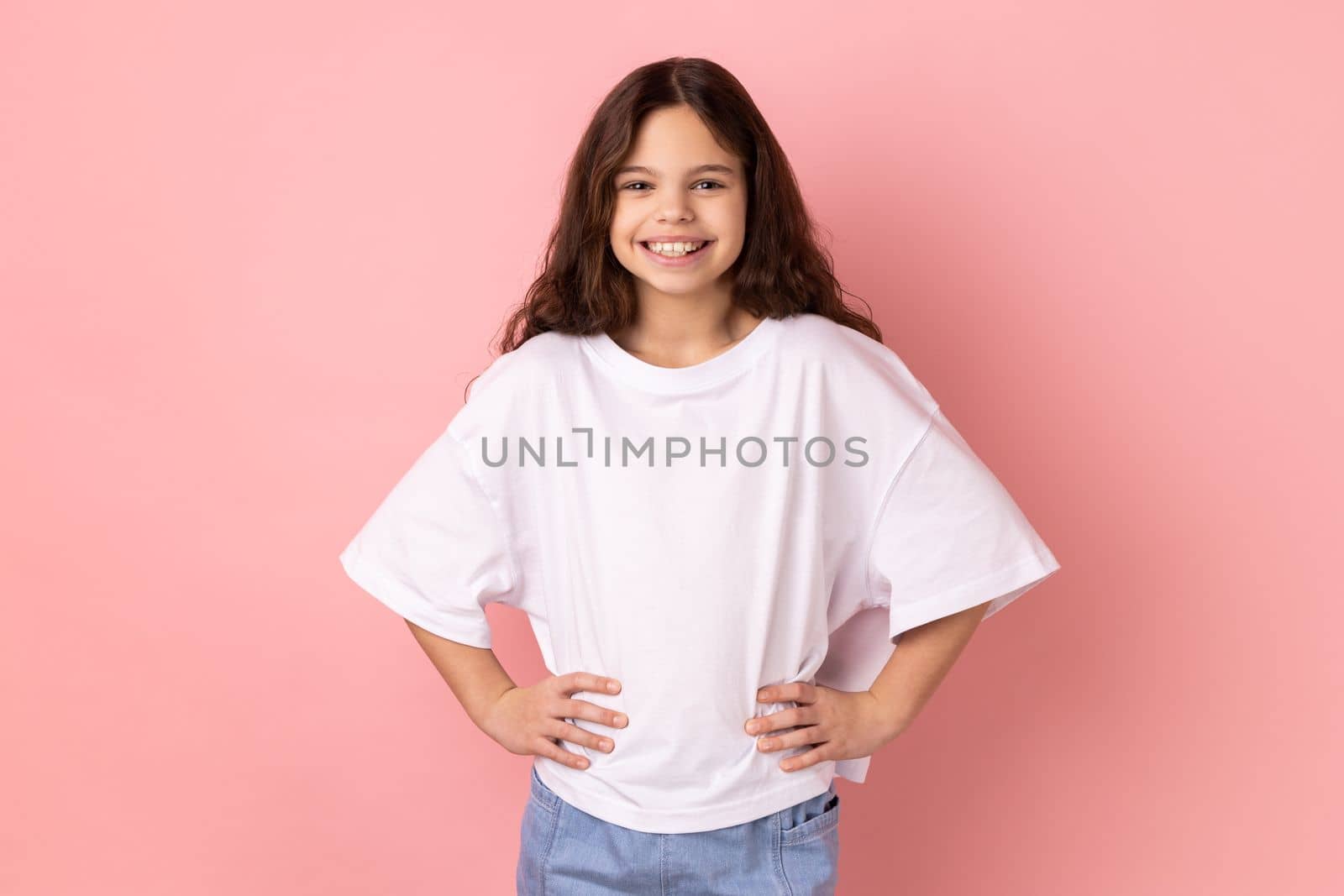 Portrait of delighted smiling little girl wearing white T-shirt standing hands on hips, looking at camera with happy expression. Indoor studio shot isolated on pink background.