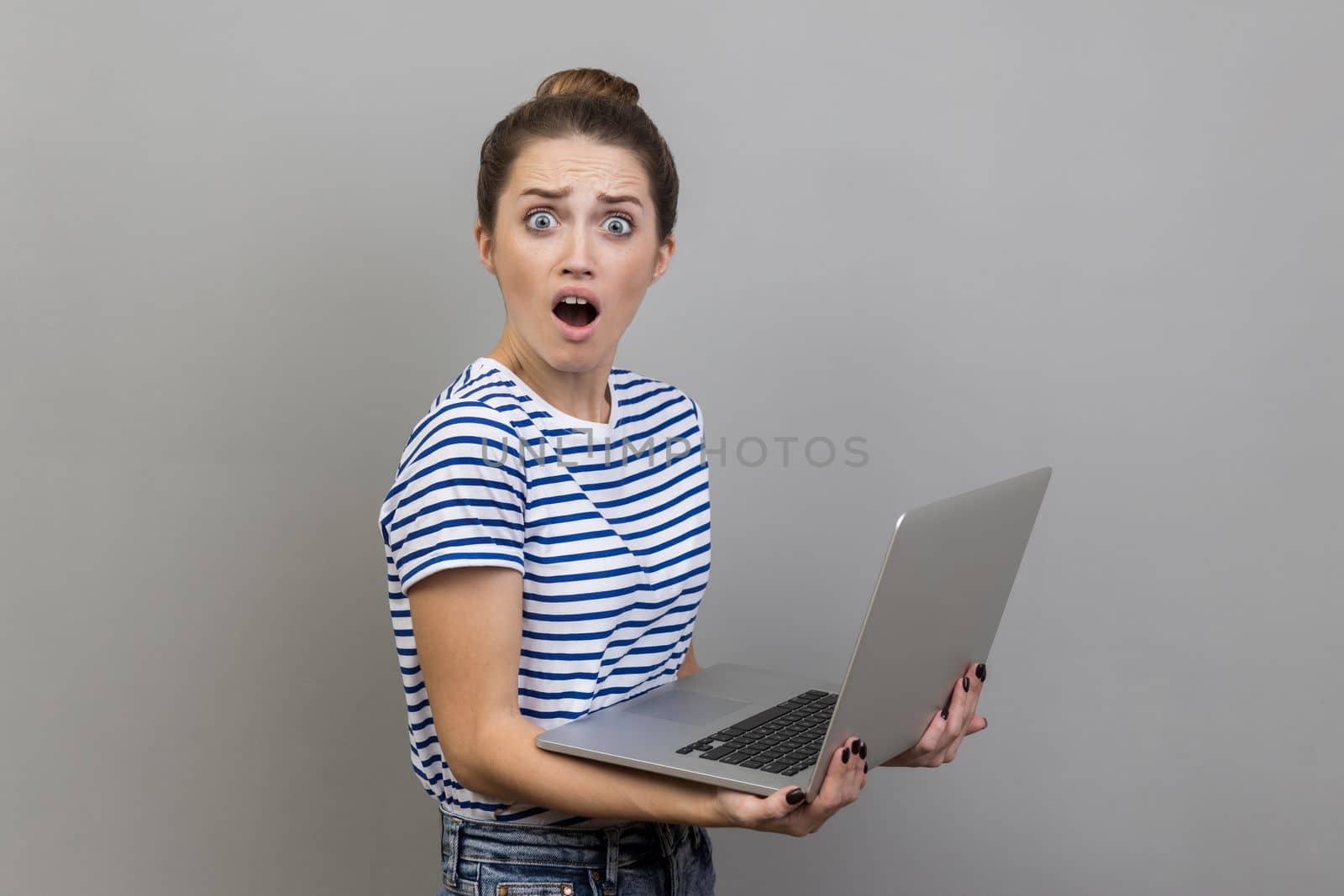 Side view of shocked woman wearing striped T-shirt working on laptop with surprised expression, freelance work, looking at camera with open mouth. Indoor studio shot isolated on gray background.