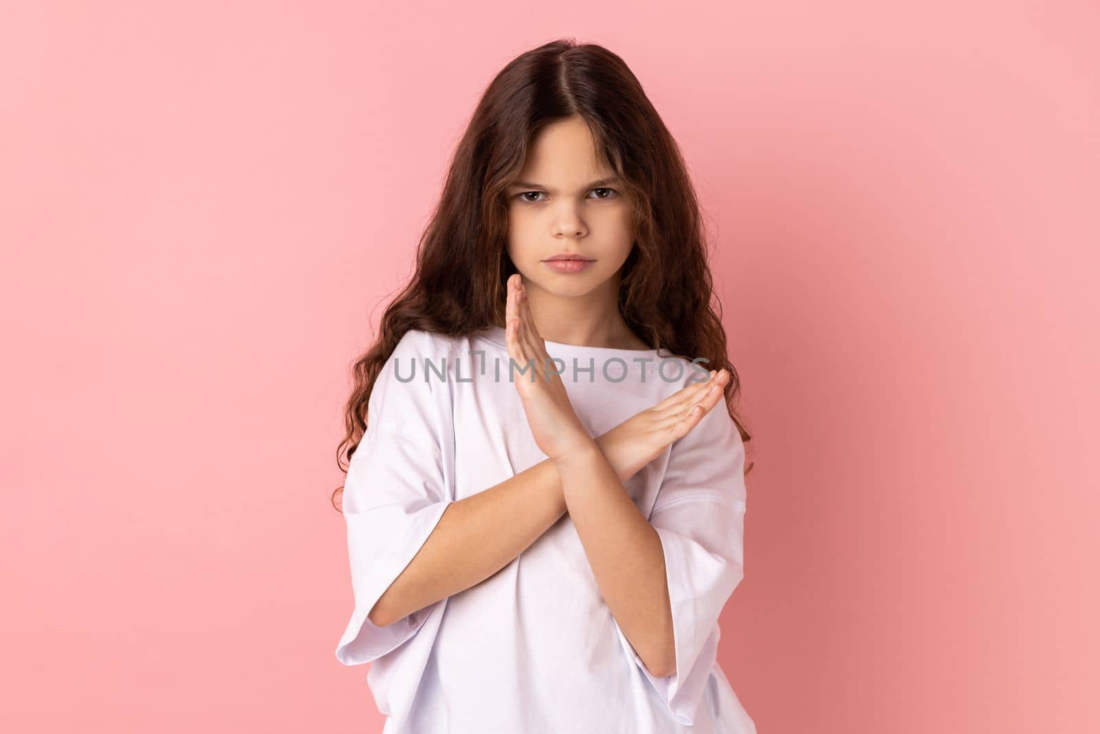 No way, absolutely not. Portrait of little girl wearing white T-shirt showing x sign with crossed hands, meaning stop, this is the end. Indoor studio shot isolated on pink background.