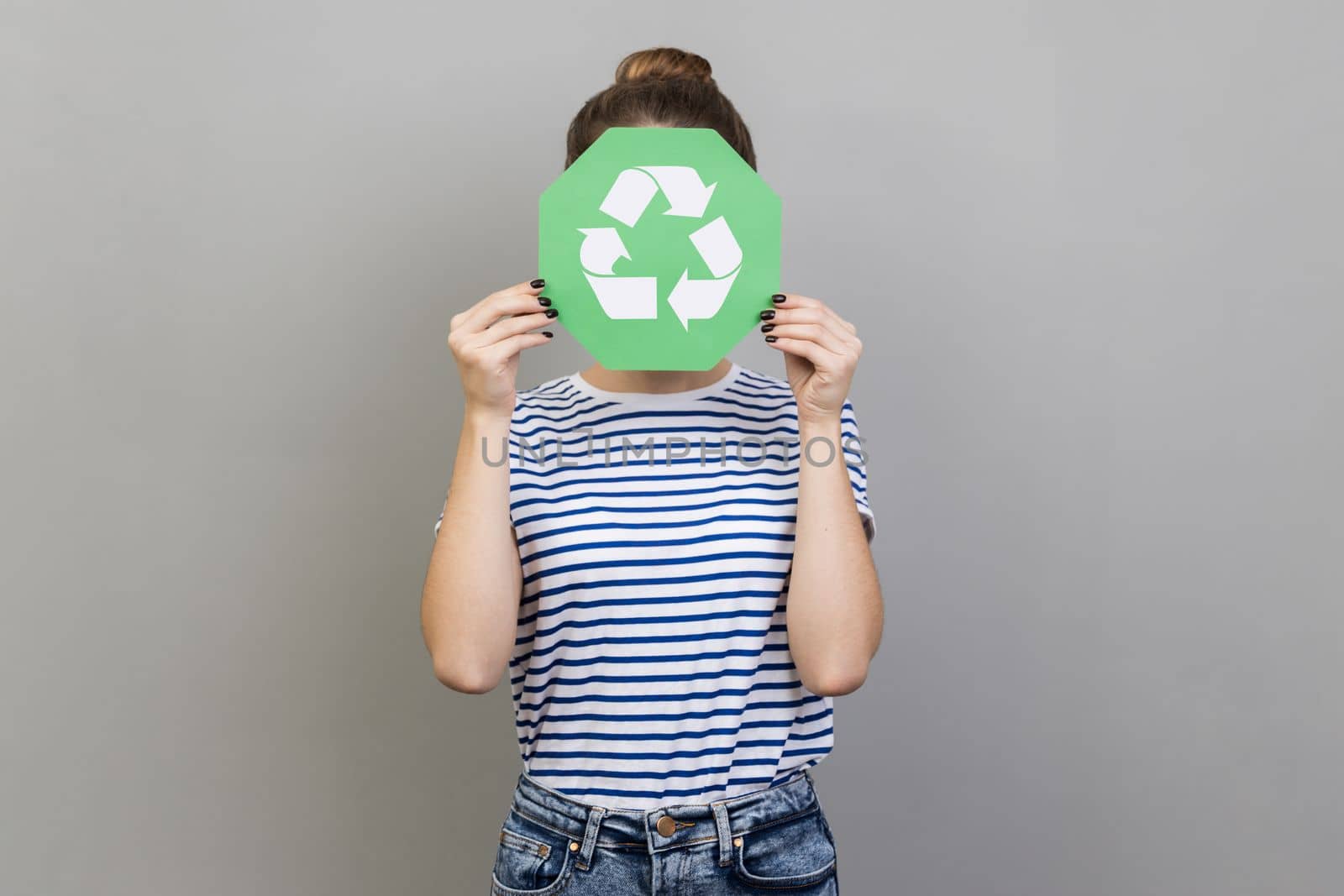 Portrait of anonymous unknown woman wearing striped T-shirt covering her face with green recycling sign, environmental problems. Indoor studio shot isolated on gray background.