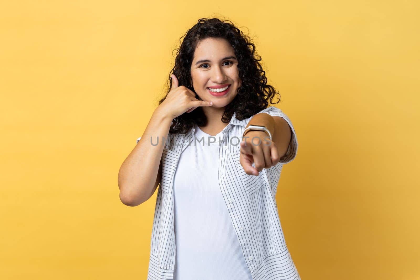 Hey you, call me back, let's contact by phone. Portrait of woman with dark wavy hair standing pointing to camera and showing call me gesture. Indoor studio shot isolated on yellow background.