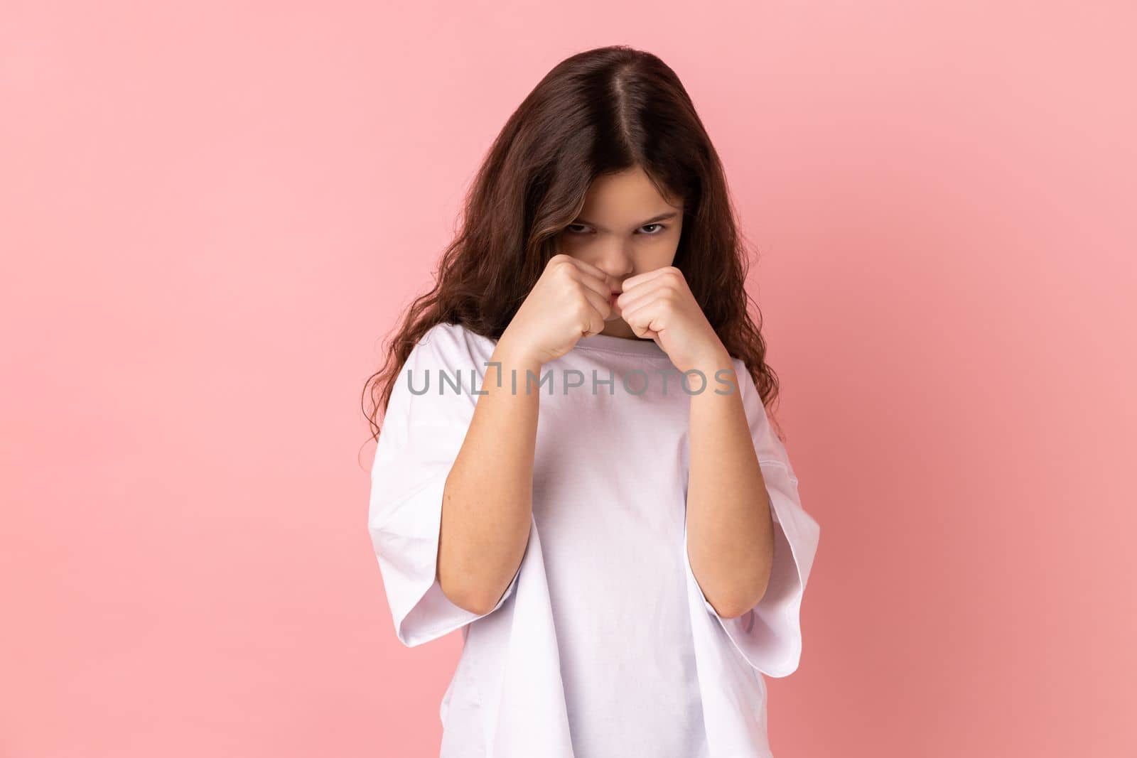 Portrait of serious concerned little girl wearing white T-shirt holding clenched fists up ready to boxing, kids self defense courses, fight spirit. Indoor studio shot isolated on pink background.