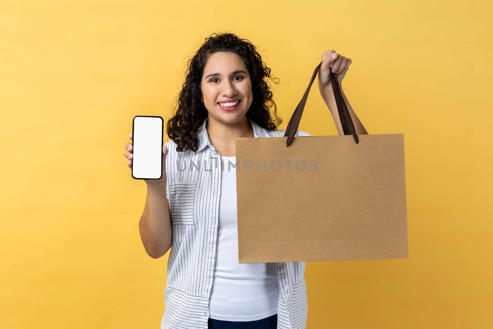 Portrait of smiling happy woman with dark wavy hair holding paper shopping bag and smart phone with empty display for advertisement. Indoor studio shot isolated on yellow background.
