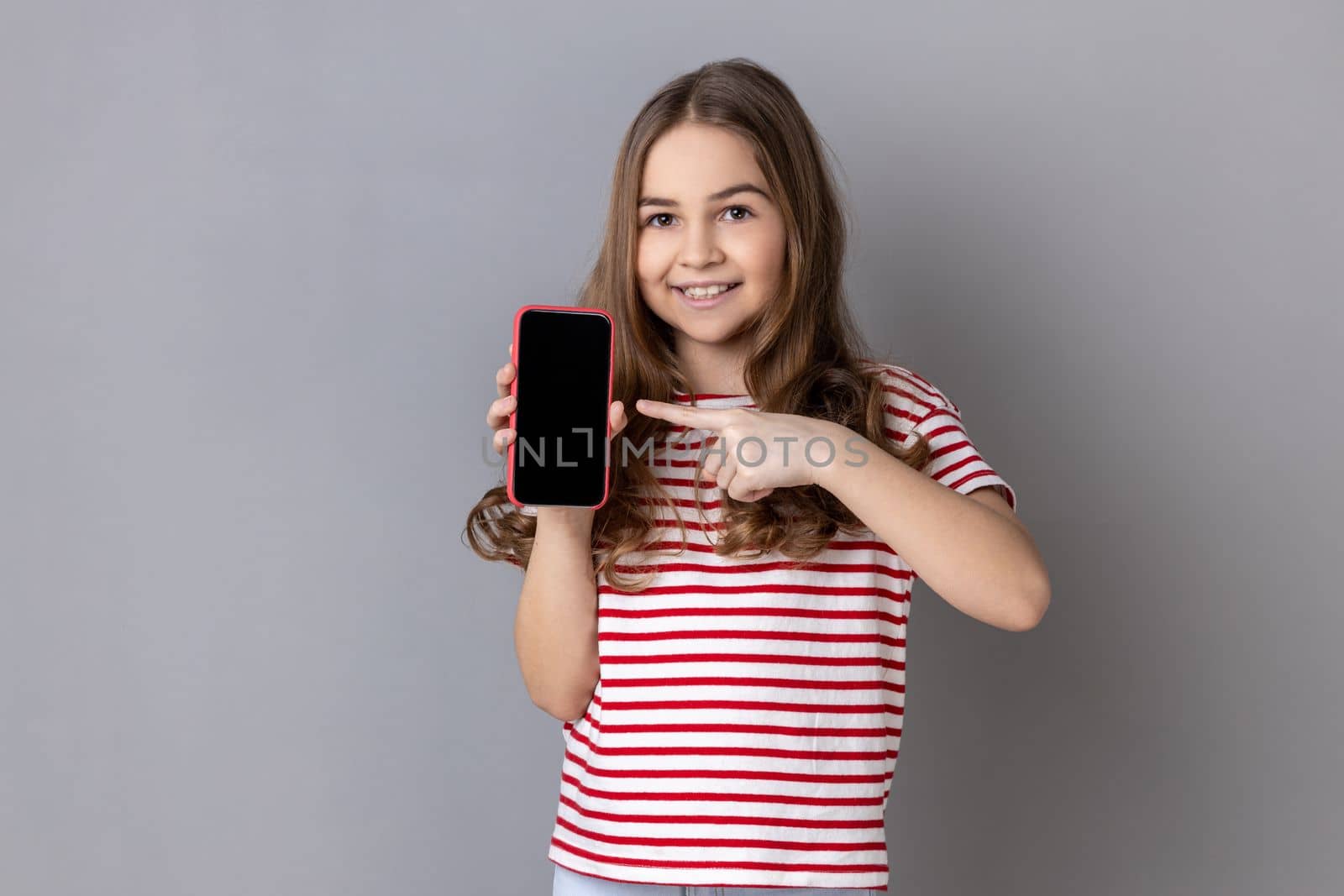 Portrait of delighted little girl wearing striped T-shirt pointing finger at smartphone with white empty display, bragging with application. Indoor studio shot isolated on gray background.