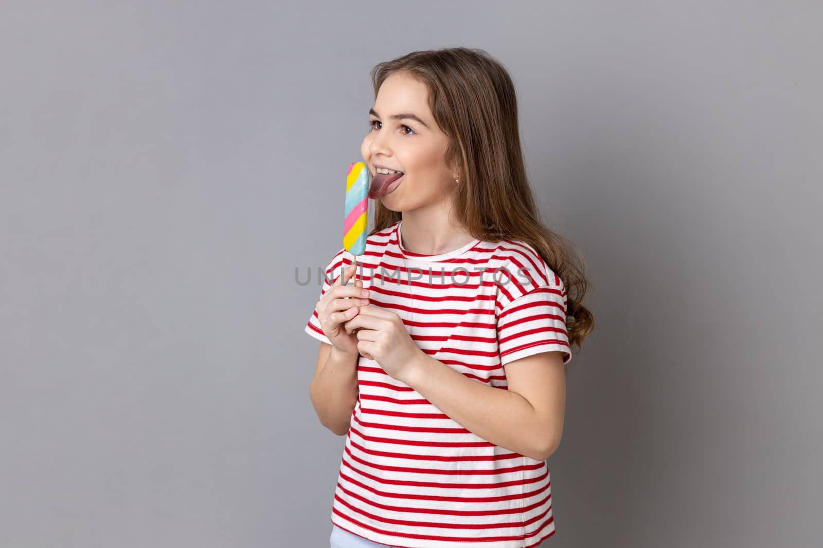 Portrait of little girl wearing striped T-shirt holding colorful candy, licking, looking away with childish satisfied expression. Indoor studio shot isolated on gray background.