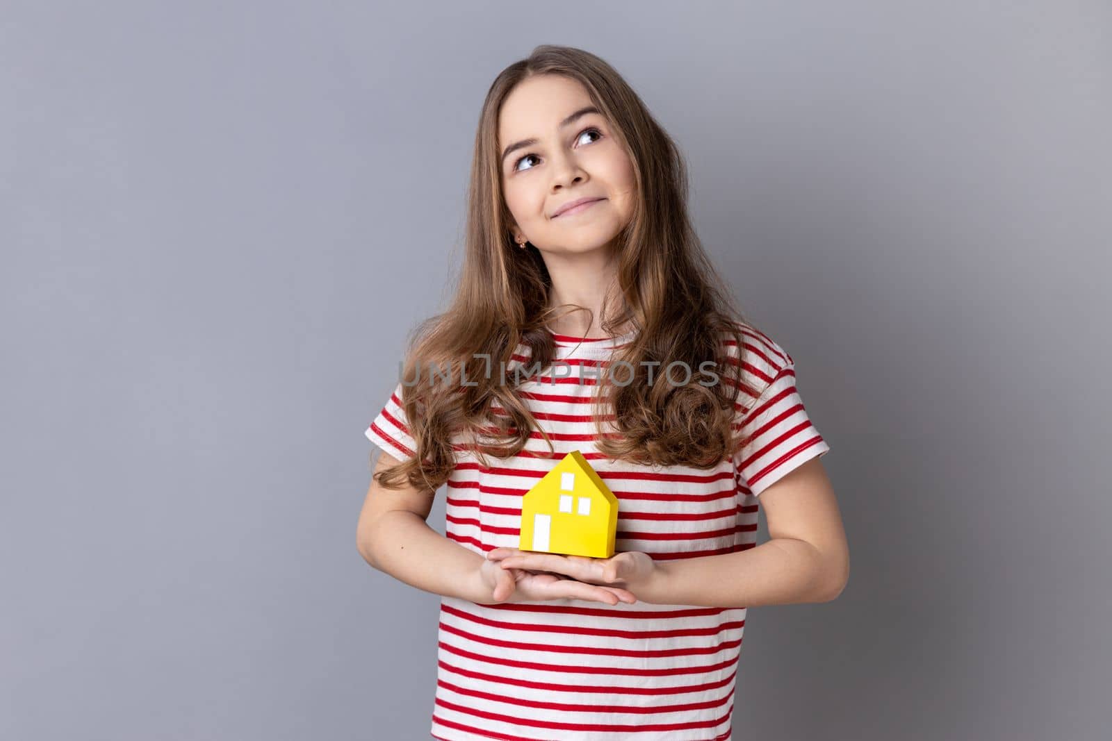 Portrait of little girl wearing striped T-shirt holding in hands little paper house looking away with dreamy expression, new apartment. Indoor studio shot isolated on gray background.