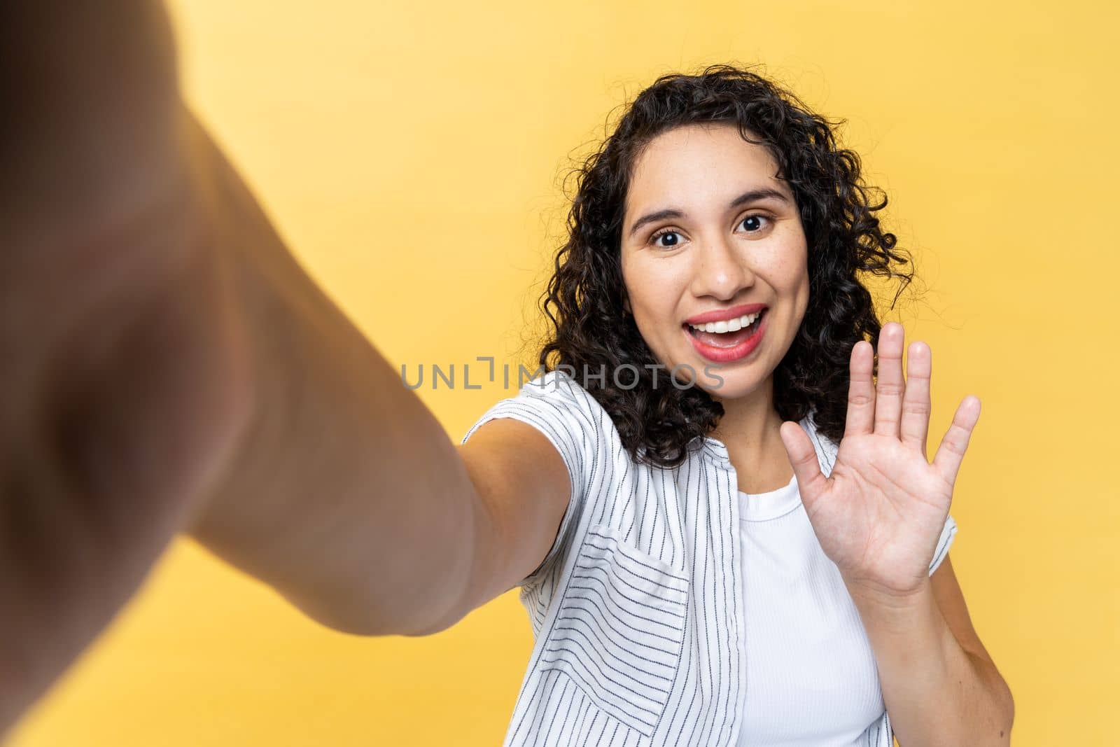 Portrait of smiling friendly woman with dark wavy hair making point of view photo, waving hand, saying hello or good bye, POV. Indoor studio shot isolated on yellow background.