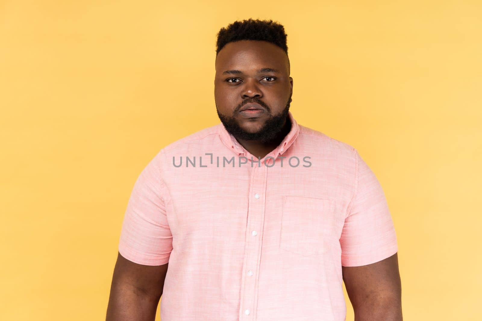 Portrait of serious strict young adult man wearing pink shirt standing and looking at camera with bossy facial expression, being in bad mood. Indoor studio shot isolated on yellow background.