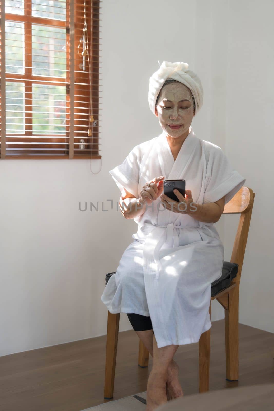 Beautiful mature Asian lady in bathrobe with facial moisturizing or anti aging clay mask on her face is using her mobile phone in the room. by wichayada