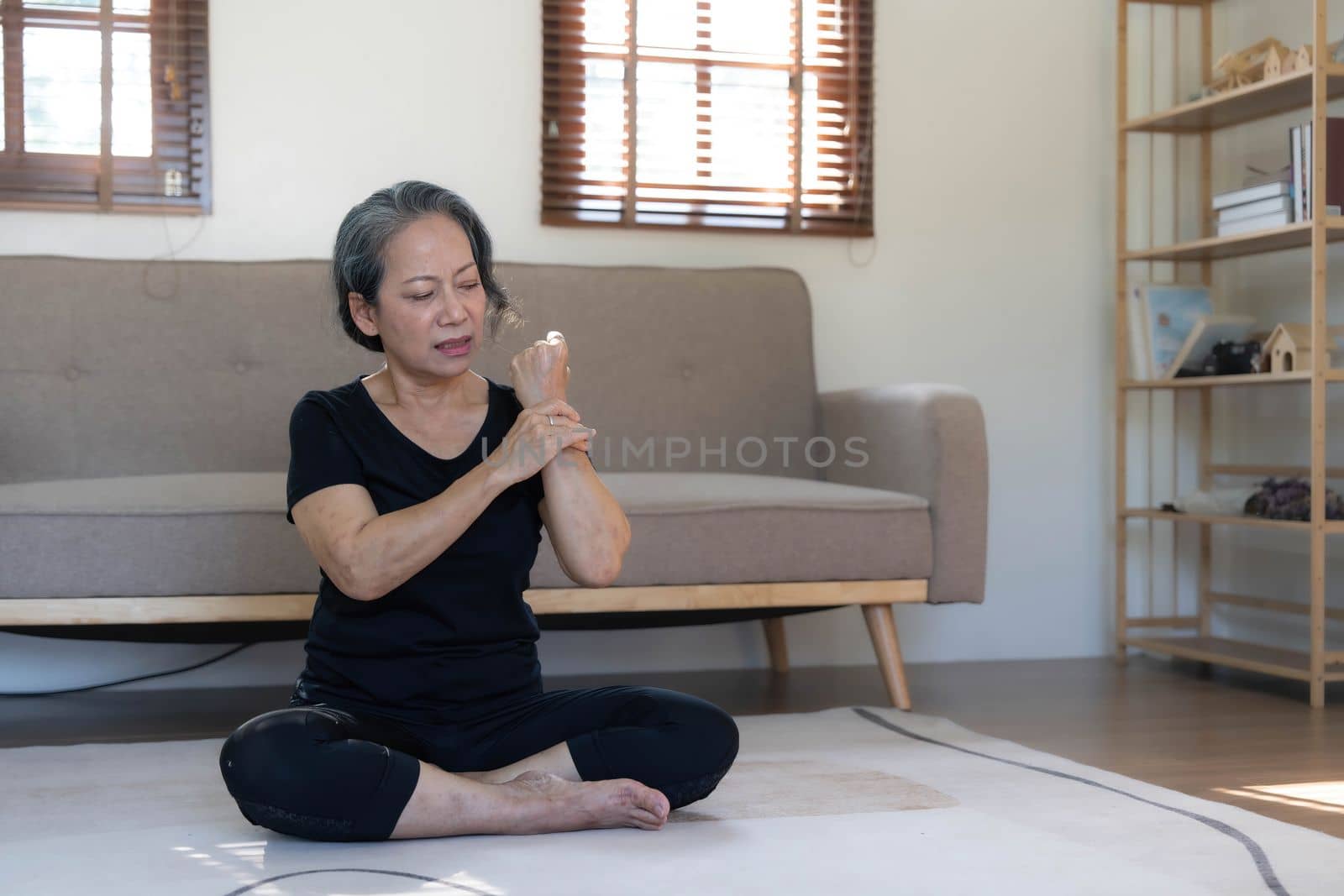 60s aged Asian woman massaging her wrist, feeling pain and swelling in the joints, injured hand during yoga practice at home. by wichayada