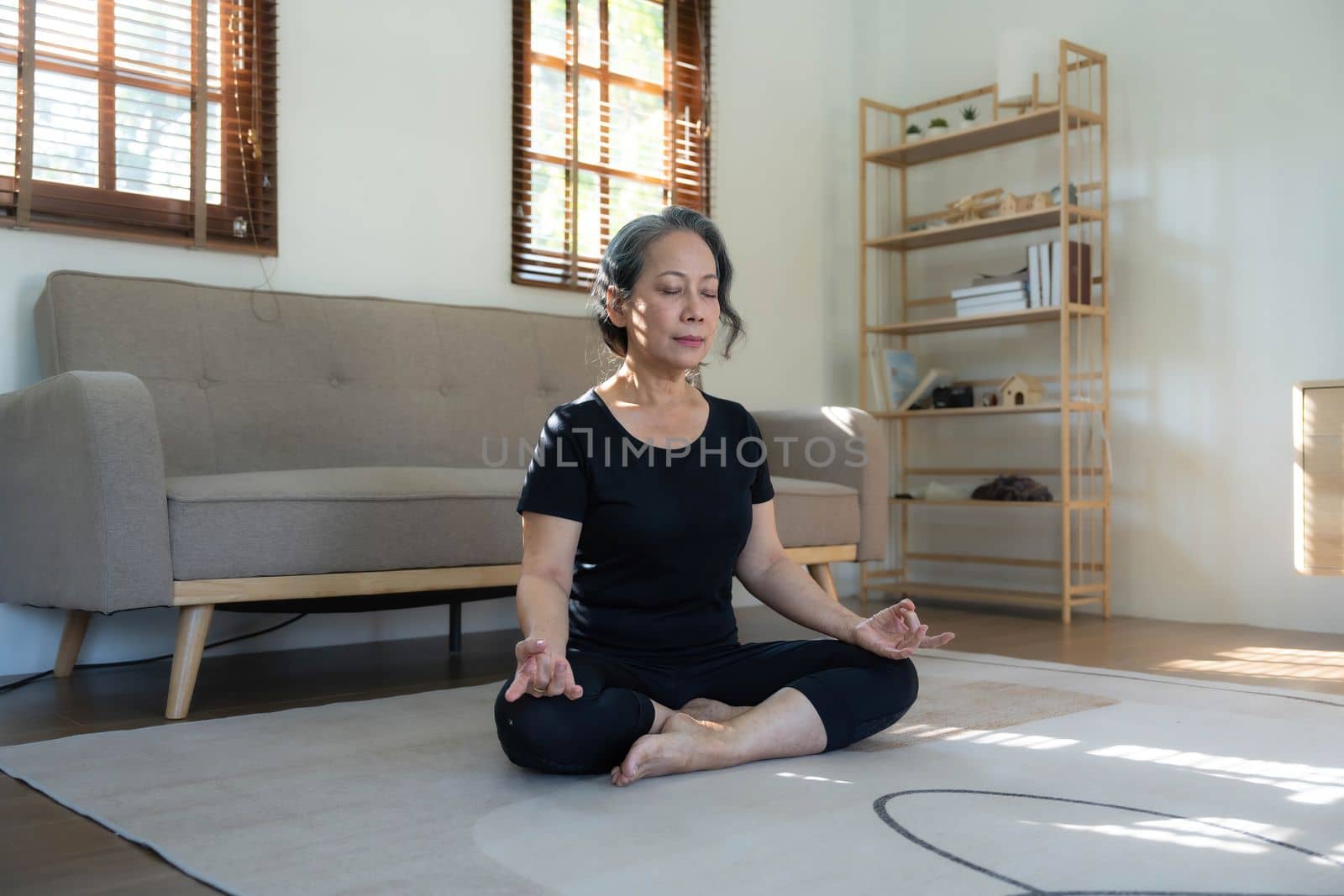 Calm and relaxed 60s retired Asian woman in gym clothes is meditating in her living room, eyes closed, practicing lotus yoga pose at home. by wichayada