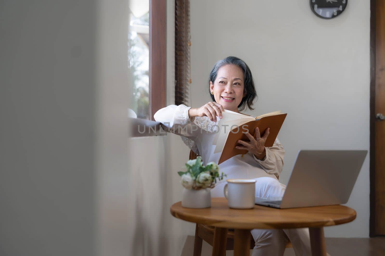 Relaxed and calm 60s retired Asian woman looking out the window, daydreaming while reading a book in her home living room. lifestyle concept by wichayada