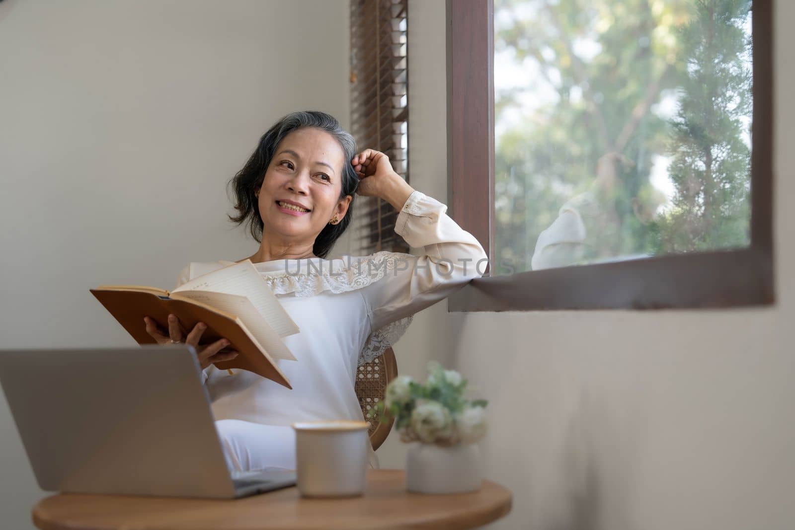 Relaxed and calm 60s retired Asian woman looking out the window, daydreaming while reading a book in her home living room. lifestyle concept by wichayada