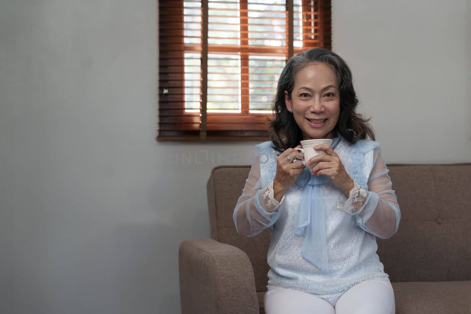 Portrait, Relaxed and calm 60s aged Asian woman sipping hot tea, having an afternoon tea in her living room. lifestyle concept by wichayada