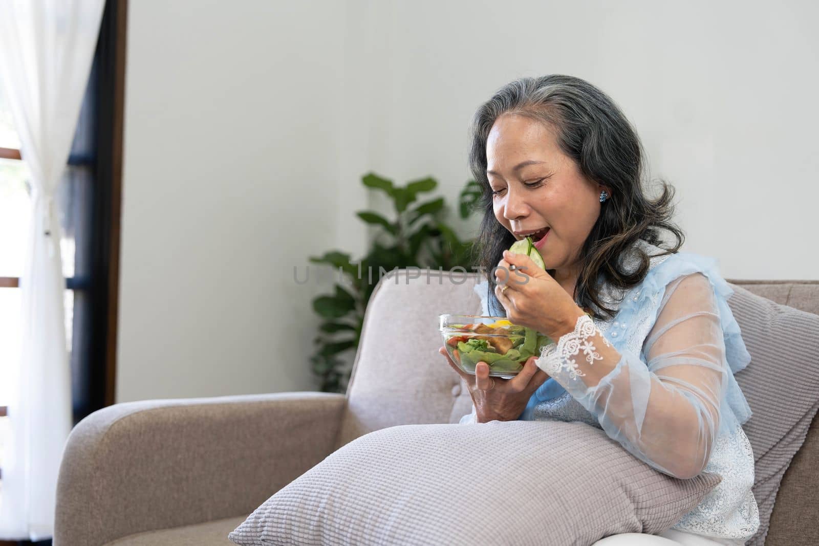 Portrait, Cheerful Asian 60s aged woman having breakfast in her living room, eating healthy salad vegetables mix bowl. by wichayada