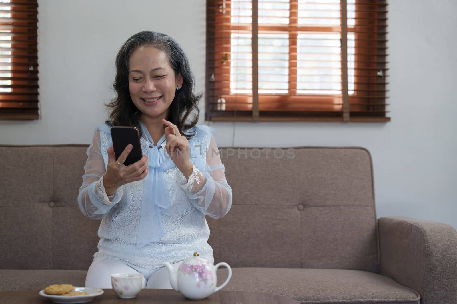 Cheerful 60s retired Asian woman using her mobile phone to chat with her grandchild while relaxing in her living room...