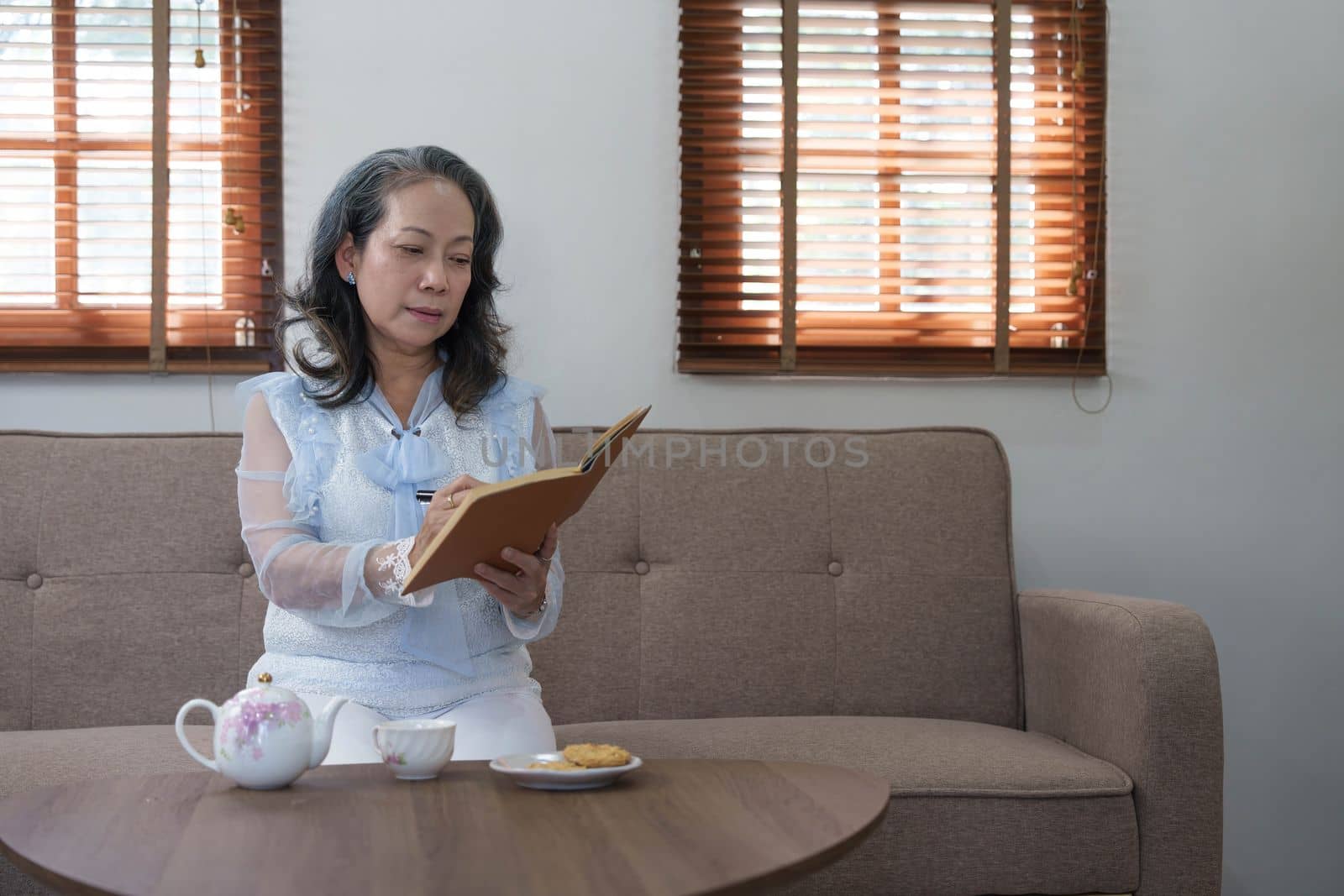 Portrait, Gorgeous 60s aged Asian woman in casual clothes eating cookies and writing her diary while relaxing in her living room by wichayada