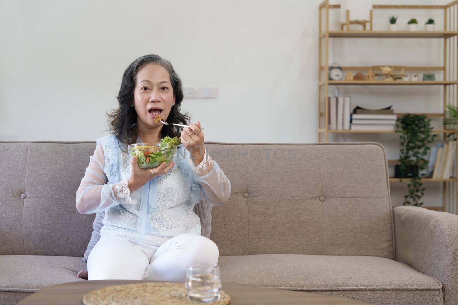 Portrait, Cheerful Asian 60s aged woman having breakfast in her living room, eating healthy salad vegetables mix bowl...