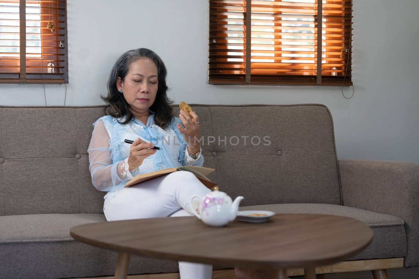 Portrait, Gorgeous 60s aged Asian woman in casual clothes eating cookies and writing her diary while relaxing in her living room by wichayada