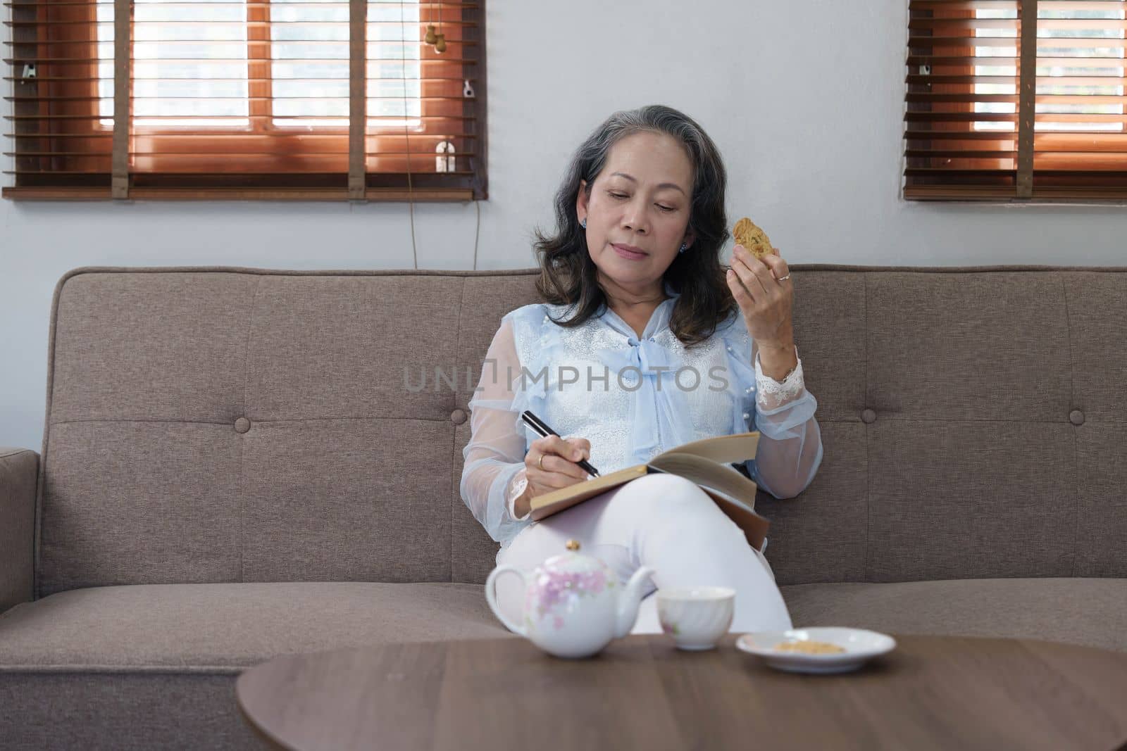 Portrait, Gorgeous 60s aged Asian woman in casual clothes eating cookies and writing her diary while relaxing in her living room by wichayada