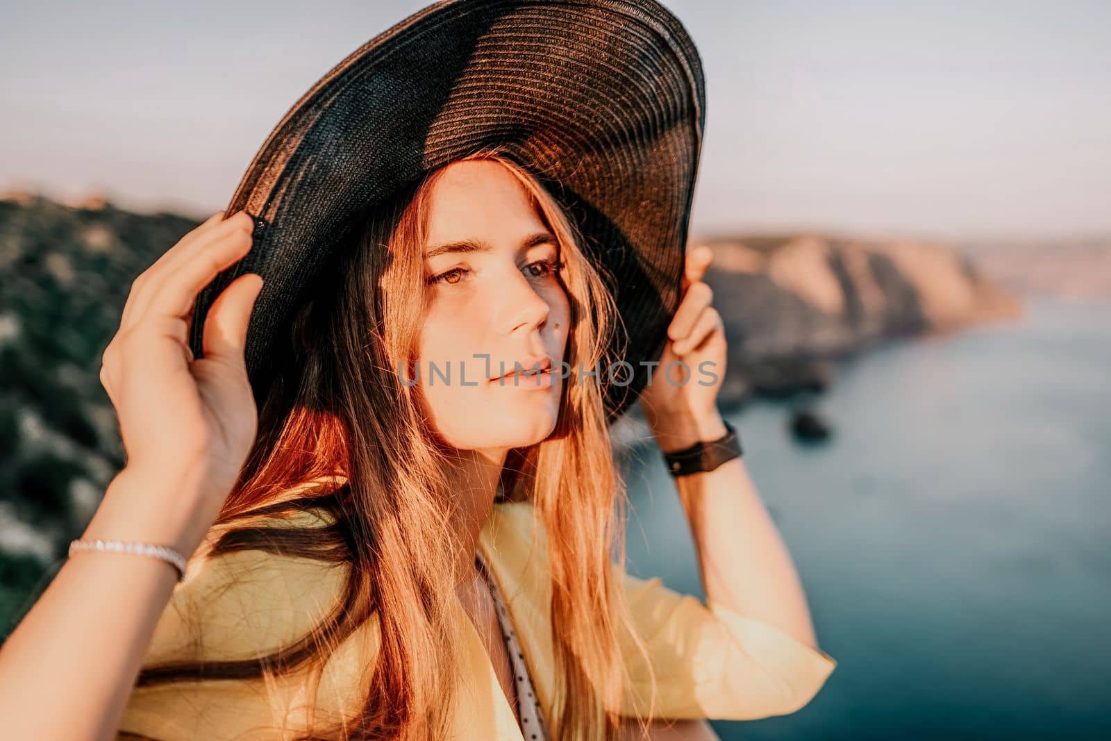 Portrait of happy young woman wearing summer black hat with large brim at beach on sunset. Closeup face of attractive girl with black straw hat. Happy young woman smiling and looking at camera at sea