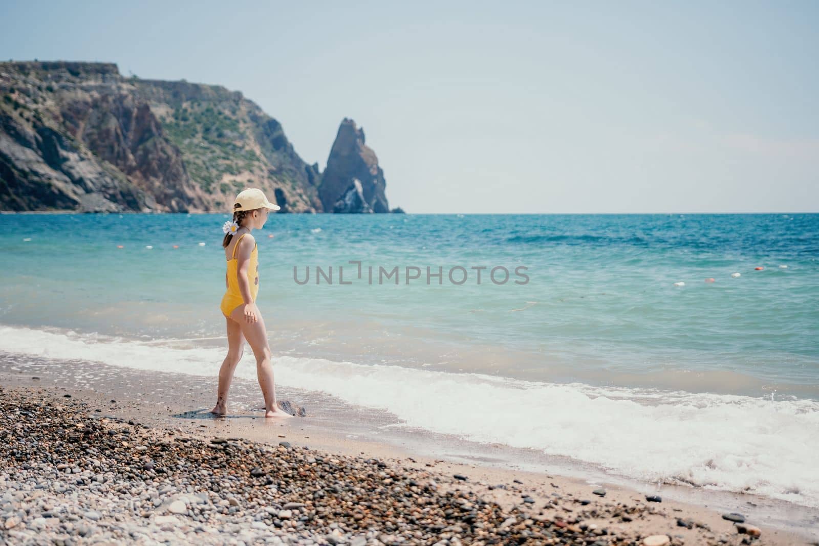 Cute little girl running along the seashore against a clear blue sea and rejoices in the rays of the summer sun. Beautiful girl in yellow swimsuit running and having fun on tropical beach