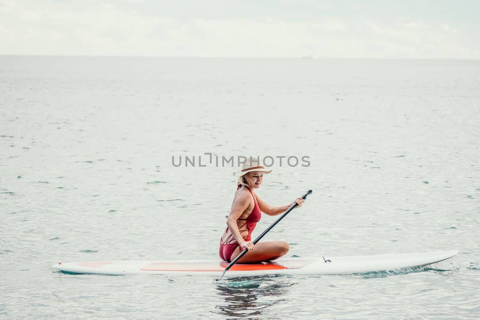 Woman in red bikini on sup board. Happy lady with blond hair in red bathing suit chilling and sunbathing by turquoise sea ocean on sunset. Holiday, vacation and recreational concept. by panophotograph