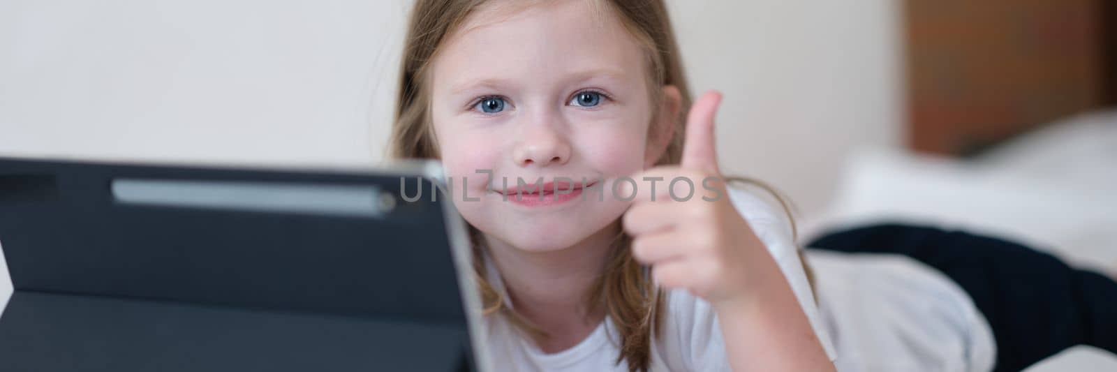 Llittle girl is lying on bed with tablet and holding thumbs up by kuprevich