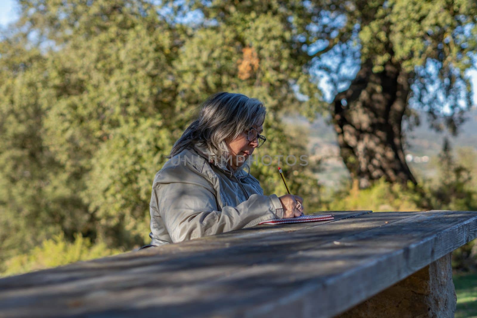 white-haired woman with glasses taking notes in her notebook in the park on a sunny winter's day