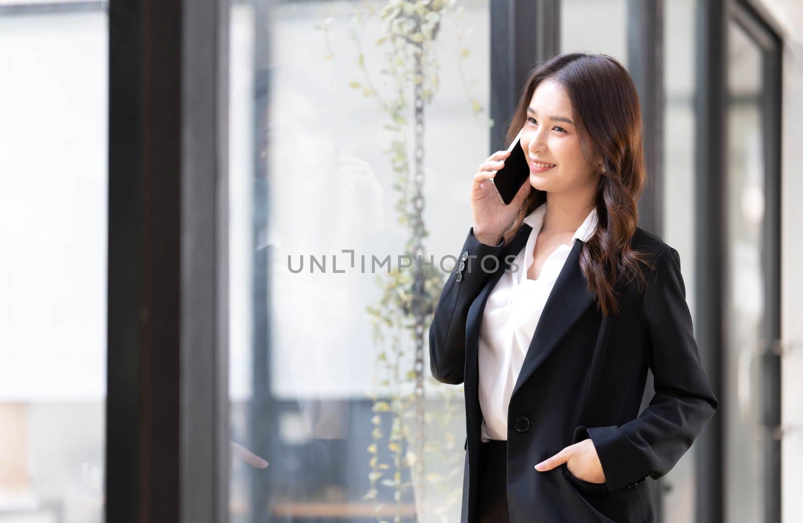 Happy young asian business woman wearing suit holding mobile phone standing in her workstation office..