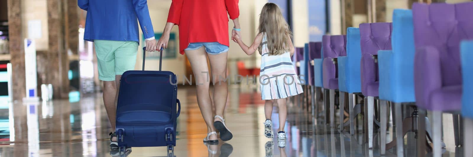 Rear view of family with suitcase while walking in airport or hotel. Parents and little daughter holding hands walking with luggage to boarding gate concept